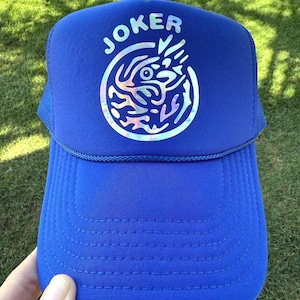 May include: A royal blue trucker hat with the word "JOKER" in white letters above a circular, iridescent graphic. The hat has a mesh back and a curved brim, suitable for sun protection.