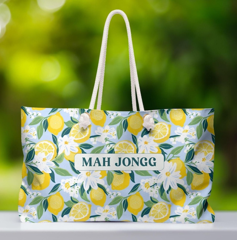 Pu&ograve; includere: Una grande borsa tote con sfondo azzurro chiaro con un motivo di limoni gialli, fiori bianchi e foglie verdi. La borsa ha manici in corda bianca e la scritta "MAH JONGG" sul davanti.