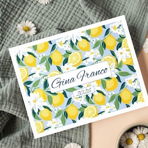 Può includere: Una carta rettangolare con un motivo a limoni, con limoni gialli, fiori bianchi e foglie verdi su sfondo azzurro. La carta riporta il nome "Gina Franco" e l'anno "2026".