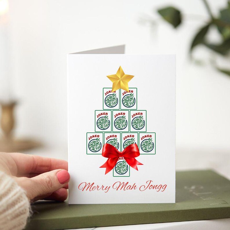 May include: A white greeting card with a Christmas tree design made of playing cards. The tree is topped with a gold star and a red bow. The text "Merry Mah Jongg" is written in red at the bottom.