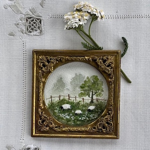 Pode incluir: Uma pequena pintura em aquarela de ovelhas pastando em um campo, emoldurada em uma moldura dourada ornamentada. A pintura apresenta um campo verde, uma cerca de madeira e uma árvore. Flores brancas e vegetação são visíveis. Um raminho de flores brancas repousa sobre a moldura.