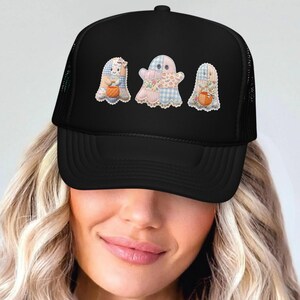 May include: A black trucker hat featuring a graphic print of three ghosts with patchwork patterns in shades of blue, pink, and cream. The ghosts are depicted holding small orange pumpkins, and the hat has a structured front panel and mesh back.