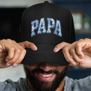 May include: A black trucker hat featuring the word PAPA in a blue and white checkered pattern with floral accents. The cap has a structured front panel and a curved brim, with the text displayed in a bold, arched, collegiate-style font.
