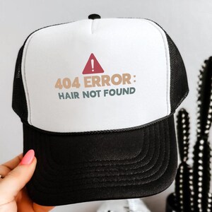May include: A black and white trucker hat with the text "404 ERROR: HAIR NOT FOUND" in brown and green. The hat has a white front panel and a black mesh back. A red triangle with an exclamation point is above the text.