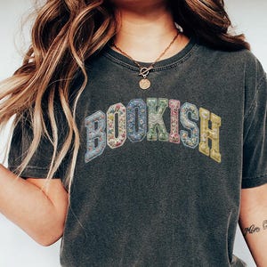 May include: Dark gray t-shirt with the word "BOOKISH" in colorful, floral-patterned letters. The shirt has a vintage, worn look. The model is wearing a gold necklace and bracelets.