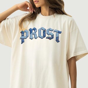 May include: An ivory-colored short-sleeve t-shirt featuring the word PROST printed across the chest in a blue and white checkered pattern with a floral design. The letters are styled in a bold, gothic-inspired font with a dark blue outline.