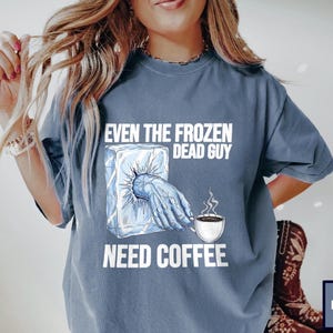 May include: Denim blue t-shirt with the text "EVEN THE FROZEN DEAD GUY NEED COFFEE". The graphic shows a frozen hand reaching out of an ice block, holding a coffee cup with steam rising.