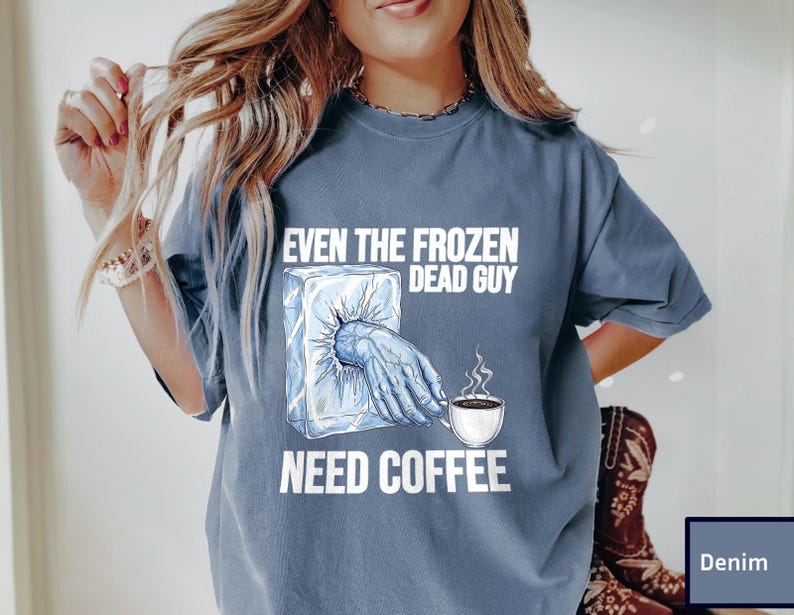May include: Denim blue t-shirt with the text "EVEN THE FROZEN DEAD GUY NEED COFFEE". The graphic shows a frozen hand reaching out of an ice block, holding a coffee cup with steam rising.
