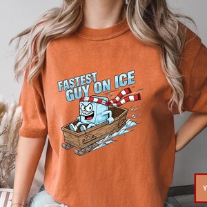 May include: Orange t-shirt with a cartoon ice cube character in a wooden sled, and the text "Fastest Guy on Ice." The ice cube wears a red and white striped scarf. The shirt is a solid orange colour.