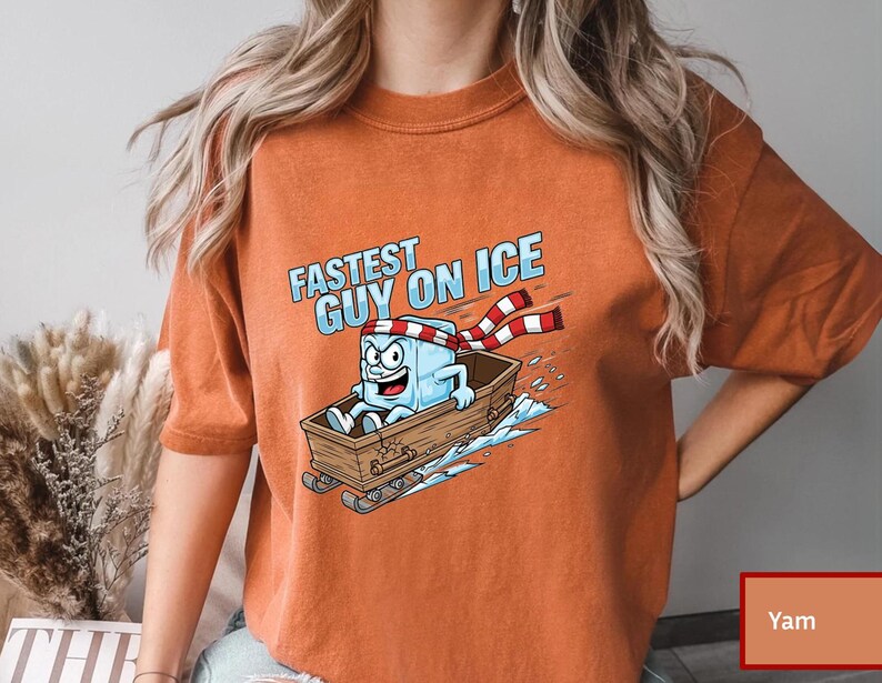 May include: Orange t-shirt with a cartoon ice cube character in a wooden sled, and the text "Fastest Guy on Ice." The ice cube wears a red and white striped scarf. The shirt is a solid orange colour.