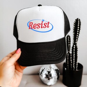 May include: Black and white trucker hat with the word "Resist" in red script inside a blue oval, with a small blue star. The hat has a mesh back and a curved brim.