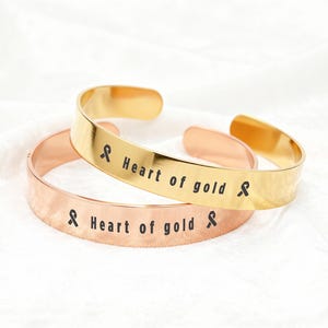 May include: Two open cuff bracelets. One is gold-colored, the other is rose gold. Both have the words "Heart of gold" and a black ribbon symbol engraved on them. The bracelets are displayed on a white surface.