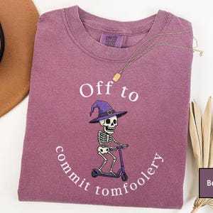 May include: A berry-coloured crewneck t-shirt featuring a graphic of a skeleton wearing a purple witch hat and riding a purple scooter. The white text reads Off to commit tomfoolery. The shirt has a Comfort Colors brand label at the inner neckline.
