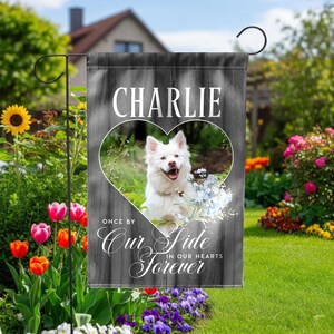 May include: A garden flag with a white dog inside a heart-shaped cutout. The flag has a gray wood-grain background and the name "Charlie" at the top. The text "Once by our side, in our hearts forever" is also on the flag.