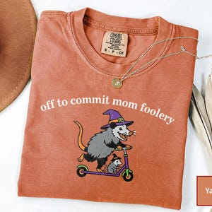 May include: A yam-coloured t-shirt featuring a graphic of an opossum wearing a purple witch hat riding a scooter with a smaller opossum, accompanied by the white text "off to commit mom foolery" printed above the illustration.