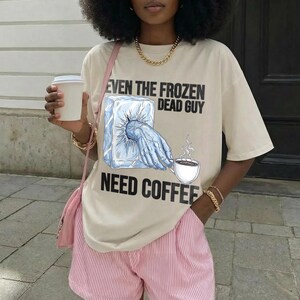 May include: Beige t-shirt with the text "EVEN THE FROZEN DEAD GUY NEED COFFEE" and a graphic of a hand reaching for a coffee cup from a block of ice. The person is wearing pink and white striped shorts and holding a white coffee cup.