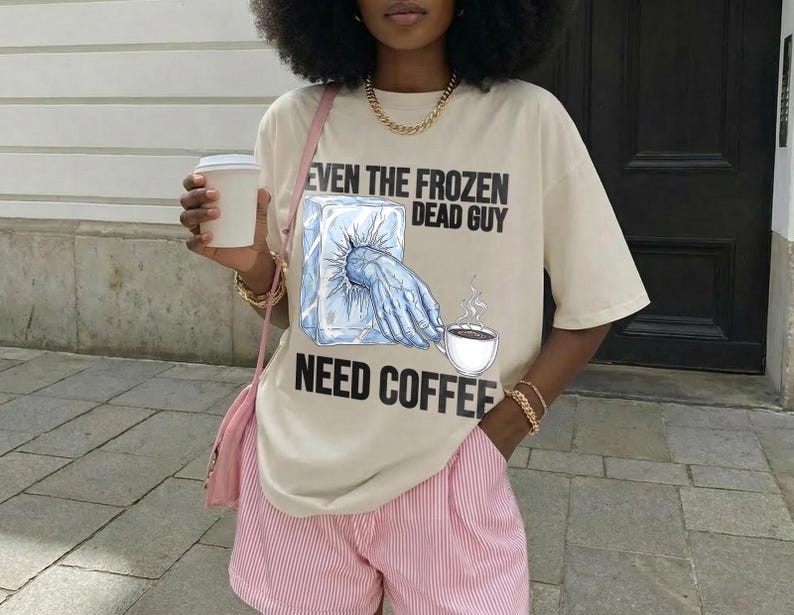 May include: Beige t-shirt with the text "EVEN THE FROZEN DEAD GUY NEED COFFEE" and a graphic of a hand reaching for a coffee cup from a block of ice. The person is wearing pink and white striped shorts and holding a white coffee cup.
