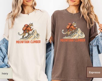 May include: Ivory and espresso-colored t-shirts featuring a cat wearing a backpack and climbing a mountain. The text "MEOWTAIN CLIMBER" is printed in orange below the cat illustration. The shirts are casual and have short sleeves.