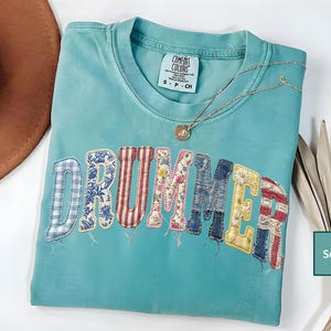 May include: A seafoam green t-shirt featuring the word DRUMMER in large, fabric-appliqued letters. Each letter is crafted from a different patterned fabric, including gingham, floral, and striped prints. The shirt has a crew neckline and a label.