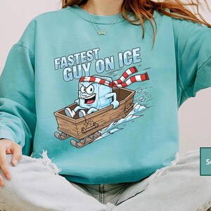 May include: Seafoam green sweatshirt featuring the text "Fastest Guy on Ice" and a cartoon ice cube character on a wooden sled. The ice cube wears a red and white striped scarf and has an angry expression.