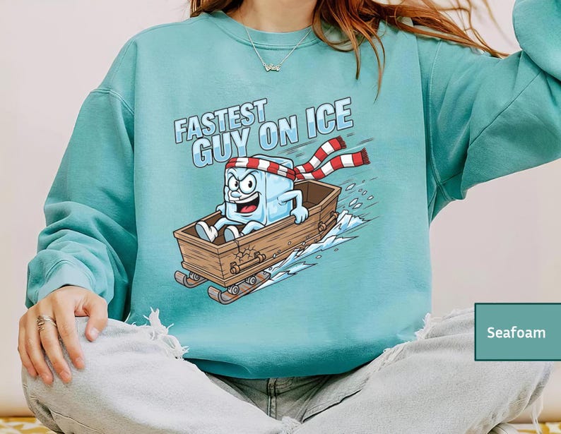 May include: Seafoam green sweatshirt featuring the text "Fastest Guy on Ice" and a cartoon ice cube character on a wooden sled. The ice cube wears a red and white striped scarf and has an angry expression.
