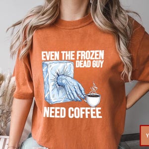 May include: Orange t-shirt with the text "EVEN THE FROZEN DEAD GUY NEED COFFEE." The graphic shows a hand reaching out of a block of ice, holding a coffee cup. The shirt is a solid orange color.