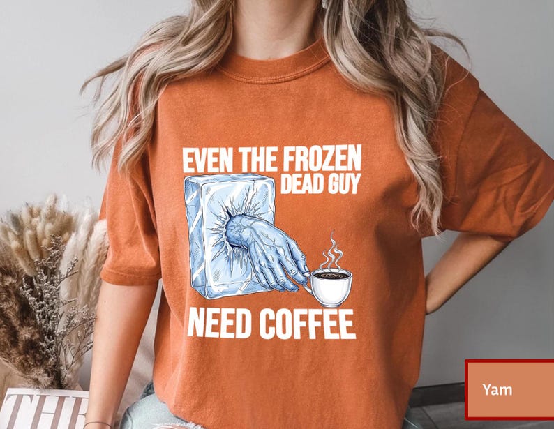 May include: Orange t-shirt with the text "EVEN THE FROZEN DEAD GUY NEED COFFEE." The graphic shows a hand reaching out of a block of ice, holding a coffee cup. The shirt is a solid orange color.