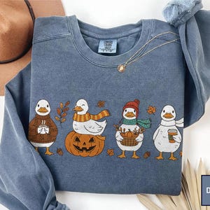 May include: A denim blue crewneck sweatshirt featuring a graphic print of four white ducks. The ducks are depicted wearing autumn-themed accessories, including a brown sweater, a striped scarf, a red beanie, and holding coffee cups and a basket.