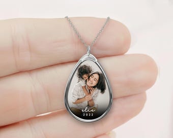 Puede incluir: Collar con colgante de lágrima plateado con una fotografía de una madre y un niño. La foto está enmarcada en un marco plateado e incluye el nombre "elie" y el año "2022" en escritura blanca. El collar se sostiene en una mano.