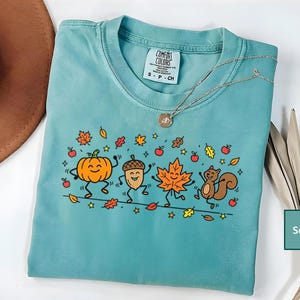 May include: A seafoam green short-sleeve t-shirt featuring a graphic print of a dancing pumpkin, acorn, maple leaf, and squirrel surrounded by small red apples and autumn leaves. The inner neck label reads Comfort Colors, 100% cotton, S-P-CH.