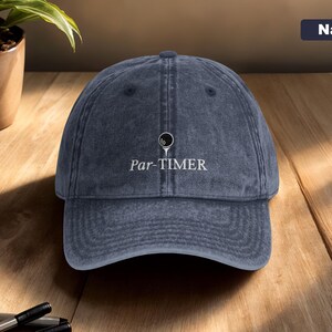 May include: Navy blue baseball cap with the text "Par-TIMER" in white. The cap features a golf ball graphic above the text. The hat is made of a soft, worn material and is sitting on a wooden surface.