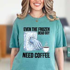 May include: Seafoam green t-shirt with the text "EVEN THE FROZEN DEAD GUY NEED COFFEE". The graphic shows a hand reaching out of a block of ice, holding a coffee cup with steam rising.