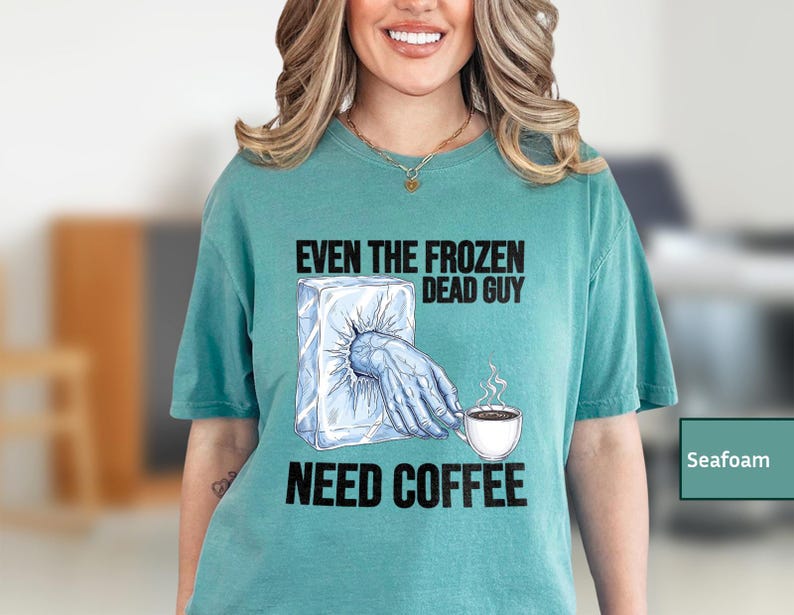 May include: Seafoam green t-shirt with the text "EVEN THE FROZEN DEAD GUY NEED COFFEE". The graphic shows a hand reaching out of a block of ice, holding a coffee cup with steam rising.