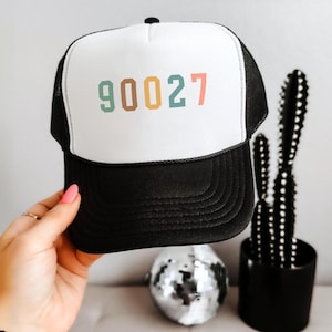 May include: A black and white trucker hat featuring a white foam front panel with the number 90027 printed in multicolored digits. The hat includes a black mesh back, a black brim with decorative stitching, and a black button on the top crown.