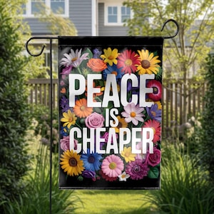May include: A black garden flag with the words "PEACE IS CHEAPER" in white, surrounded by a vibrant array of colorful flowers. The flowers include yellow sunflowers, orange gerberas, pink roses, and purple daisies. The flag is displayed outdoors.