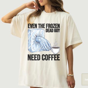 May include: Ivory-colored t-shirt featuring the text "EVEN THE FROZEN DEAD GUY NEED COFFEE." The graphic shows a hand emerging from an ice block, reaching for a steaming coffee cup. The design is in black and blue.