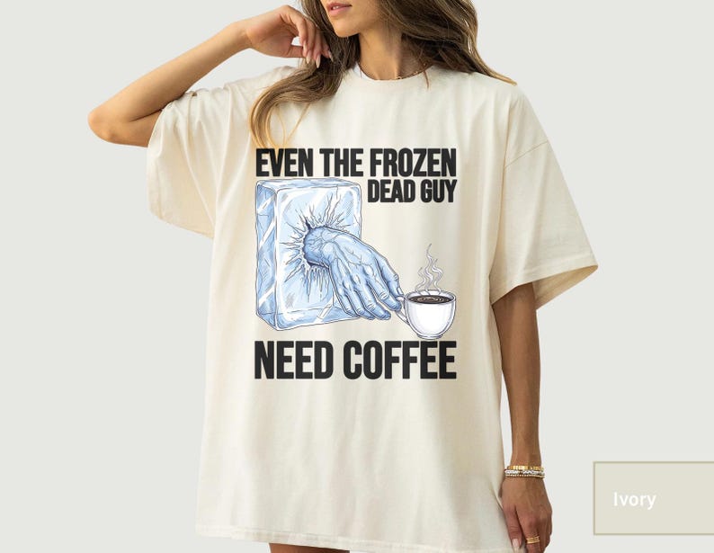 May include: Ivory-colored t-shirt featuring the text "EVEN THE FROZEN DEAD GUY NEED COFFEE." The graphic shows a hand emerging from an ice block, reaching for a steaming coffee cup. The design is in black and blue.