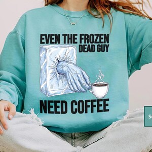 May include: Seafoam green sweatshirt with the text "EVEN THE FROZEN DEAD GUY NEED COFFEE." The graphic shows a hand emerging from a block of ice, reaching for a steaming cup of coffee. The sweatshirt is a comfortable, casual style.