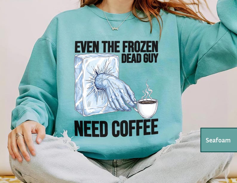 May include: Seafoam green sweatshirt with the text "EVEN THE FROZEN DEAD GUY NEED COFFEE." The graphic shows a hand emerging from a block of ice, reaching for a steaming cup of coffee. The sweatshirt is a comfortable, casual style.