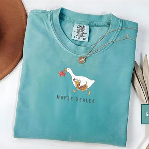 May include: Seafoam green t-shirt with a graphic of a white goose carrying a maple leaf and a briefcase. The text "MAPLE DEALER" is printed below the goose. A silver necklace with a pendant is on the shirt.