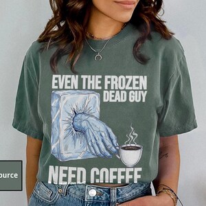May include: Blue Spruce t-shirt with the text "EVEN THE FROZEN DEAD GUY NEED COFFEE". The graphic shows a hand emerging from an ice block, holding a coffee cup. The shirt is a relaxed fit.