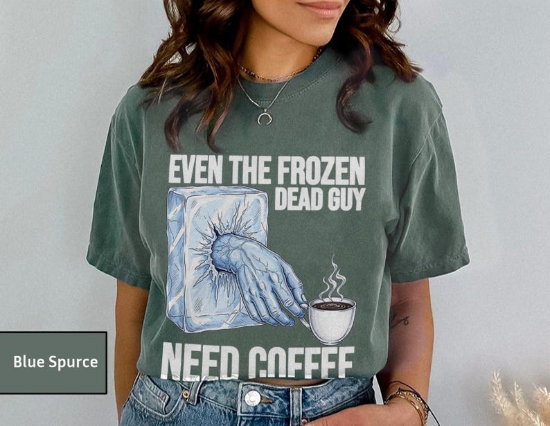 May include: Blue Spruce t-shirt with the text "EVEN THE FROZEN DEAD GUY NEED COFFEE". The graphic shows a hand emerging from an ice block, holding a coffee cup. The shirt is a relaxed fit.