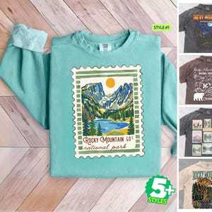 May include: A mint green crewneck sweatshirt featuring a graphic of a postage stamp with a Rocky Mountain National Park landscape, including mountains, a lake, and a sun. Five additional t-shirt styles are shown in smaller thumbnails.