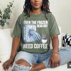 May include: Olive green t-shirt with the text "EVEN THE FROZEN DEAD GUY NEED COFFEE." The graphic shows a hand emerging from a block of ice and a coffee cup with steam. The shirt is a relaxed fit.