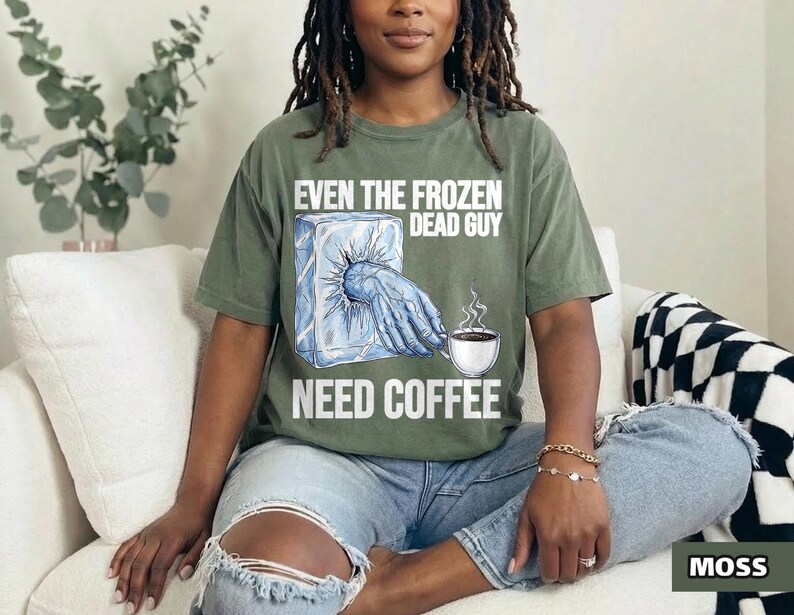 May include: Olive green t-shirt with the text "EVEN THE FROZEN DEAD GUY NEED COFFEE." The graphic shows a hand emerging from a block of ice and a coffee cup with steam. The shirt is a relaxed fit.