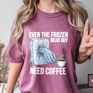 May include: Berry-colored t-shirt with the text "EVEN THE FROZEN DEAD GUY NEED COFFEE." The graphic shows a frozen hand reaching out of an ice block, holding a coffee cup with steam rising. The shirt is a casual wear item.