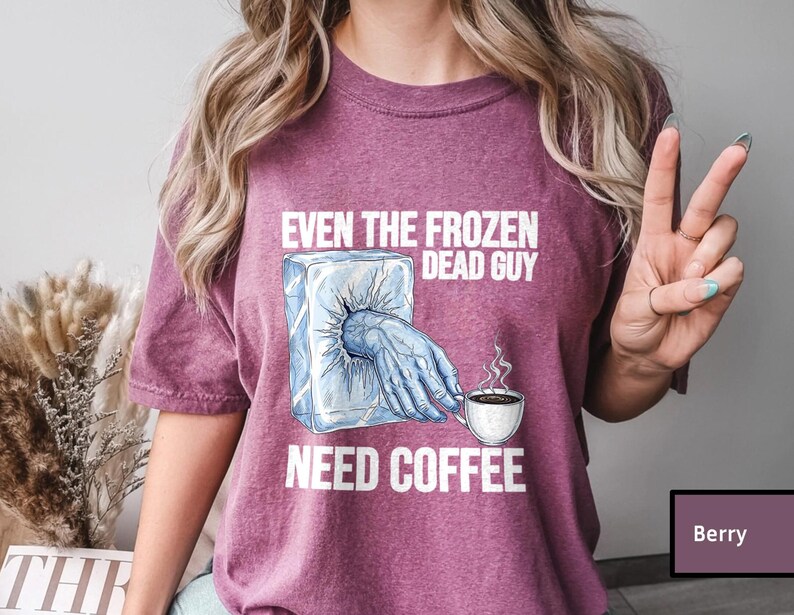 May include: Berry-colored t-shirt with the text "EVEN THE FROZEN DEAD GUY NEED COFFEE." The graphic shows a frozen hand reaching out of an ice block, holding a coffee cup with steam rising. The shirt is a casual wear item.