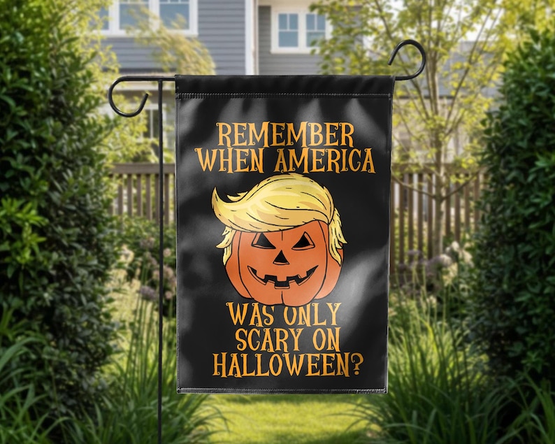 Puede incluir: Bandera de jard&iacute;n negra con el texto "REMEMBER WHEN AMERICA WAS ONLY SCARY ON HALLOWEEN?" y una calabaza con pelo rubio. La bandera est&aacute; colgada de un soporte de metal negro en un jard&iacute;n.
