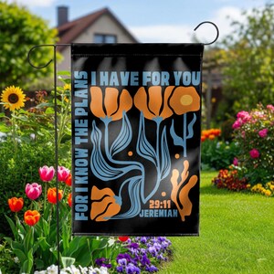 May include: A rectangular black garden flag featuring orange and light blue stylized floral illustrations. The text reads FOR I KNOW THE PLANS I HAVE FOR YOU and JEREMIAH 29:11 in light blue lettering. The flag is suspended on a black metal stand.