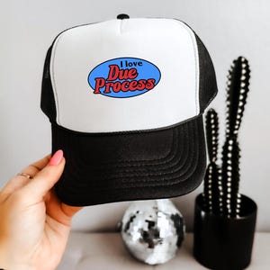 May include: A black and white trucker hat featuring a white foam front panel with a blue oval graphic containing the red text I love Due Process. The hat has a black brim and black mesh back panels with an adjustable snapback closure at the rear.